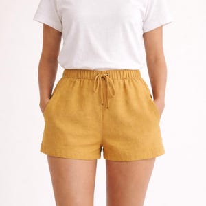 May include: A white crew neck t-shirt paired with mustard yellow shorts. The shorts have an elastic waistband with a drawstring and side pockets. The outfit is casual and comfortable.