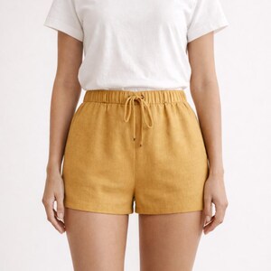 May include: A white crew neck t-shirt paired with mustard yellow shorts. The shorts have an elastic waistband with a drawstring. The outfit is casual and suitable for warm weather.