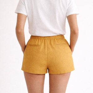 May include: Mustard yellow shorts with an elastic waistband and two back pockets. The shorts are paired with a white t-shirt. The model is facing away from the camera, with hands in the pockets.