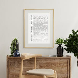 May include: A framed print with the text "Lessons in Gentility, Anne 1860" in a decorative font. The print is in a gold frame and is displayed on a wooden cabinet with decorative plants and a chair.