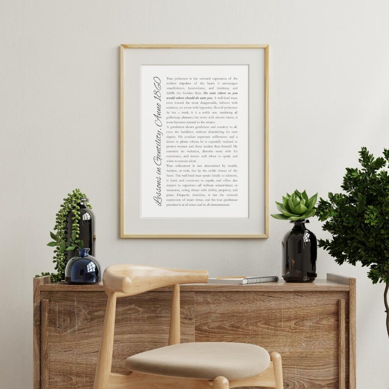 May include: A framed print with the text "Lessons in Gentility, Anne 1860" in a decorative font. The print is in a gold frame and is displayed on a wooden cabinet with decorative plants and a chair.