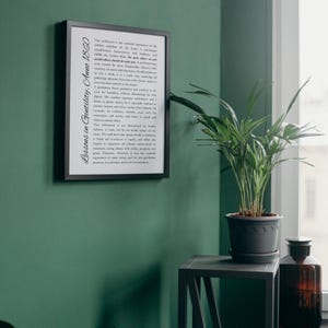 May include: A framed print with the text "Lessons in Gentility, Anno 1860" in a black frame, displayed on a dark green wall. A potted plant sits on a small gray table next to a window. A brown glass bottle is also visible.