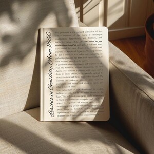 May include: A small, open book with the title "Lessons in Gentility, Anno 1860" in a serif font. The book is open to a page of text, and is resting on a beige surface. The book's pages are off-white.