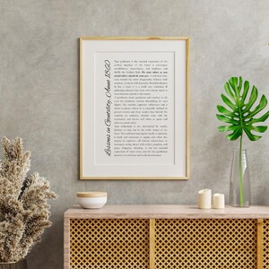 May include: A framed print with the text "Lessons in Gentility, Anno 1860" in a gold frame. The print is displayed on a wooden cabinet with a decorative front. A vase with a large green leaf and two candles are on the cabinet.