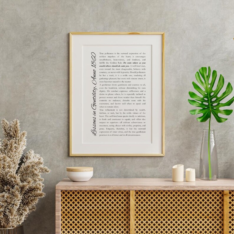 May include: A framed print with the text "Lessons in Gentility, Anno 1860" in a gold frame. The print is displayed on a wooden cabinet with a decorative front. A vase with a large green leaf and two candles are on the cabinet.
