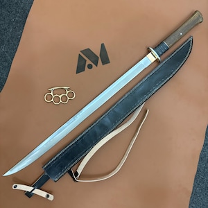 May include: A long sword with a silver blade, wooden handle, and black leather sheath. A brass knuckle duster and a logo are also present on a brown surface.