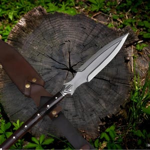 May include: A spear with a polished steel blade and a dark wooden handle with white accents. The spear is resting on a weathered tree stump, with a brown leather sheath. The spear's blade is approximately 12 inches long.
