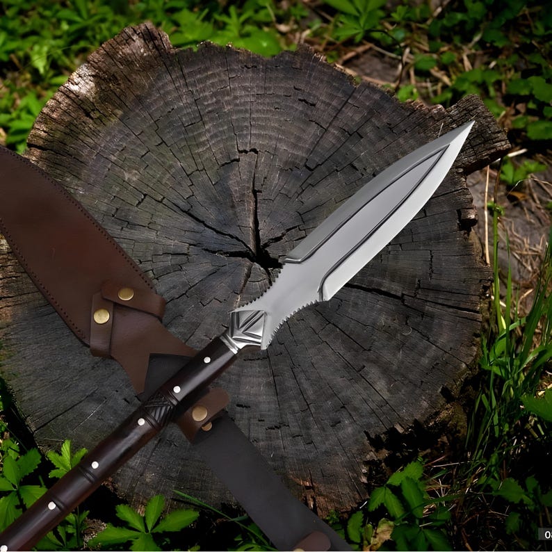 May include: A spear with a polished steel blade and a dark wooden handle with white accents. The spear is resting on a weathered tree stump, with a brown leather sheath. The spear's blade is approximately 12 inches long.