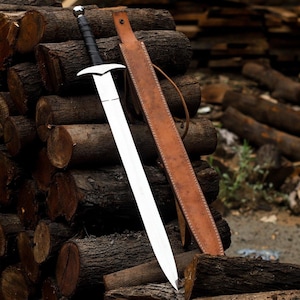 Op de afbeelding: Een middeleeuws zwaard met een zilveren lemmet en zwarte handgreep rust tegen een stapel boomstammen. Een bruine leren schede is naast het zwaard geplaatst. Het zwaard heeft een pareerstang en een zilveren pommel.