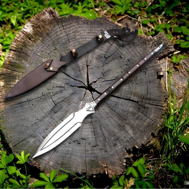 May include: A spear with a silver blade and a dark brown wooden shaft, resting on a tree stump. A brown leather sheath and strap are also visible. The spear has decorative carvings near the blade and on the shaft.