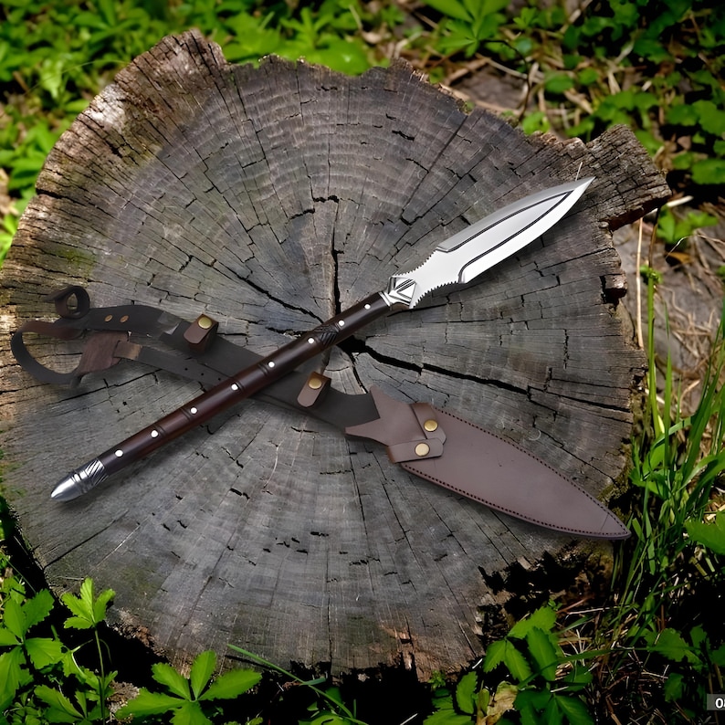 May include: A spear with a silver blade and a dark brown wooden shaft, resting on a tree stump. The spear has a leather sheath. The spearhead is silver with a decorative design near the shaft.