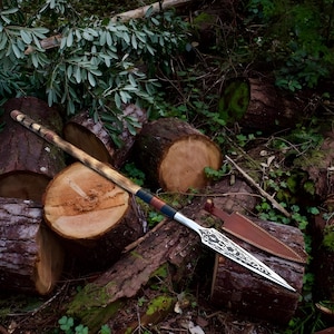 May include: A spear with a wooden shaft and a silver blade with an ornate design. The shaft has a leather-wrapped grip with brown, black, and red accents. The spear is resting on a pile of logs in a forest setting.