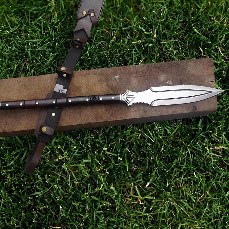 May include: A spear with a polished silver blade and a dark wooden shaft. The spear is resting on a wooden plank, with a brown leather strap and sheath visible. The spear is designed with decorative elements.