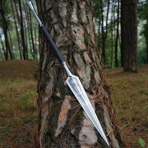 May include: A silver and black spear rests against a tree trunk in a woodland scene. The spearhead is polished silver with an ornate design. The handle is black and possibly wrapped for grip. The spear is a traditional weapon.