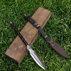 May include: A spear with a silver blade and a black handle with white accents rests on a wooden plank. A brown leather sheath and strap are next to the spear. The items are on a bed of green grass.