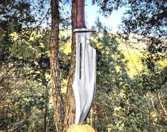 May include: A large, silver-colored knife with a wooden handle, resting on a tree stump. The blade has a unique shape with a cutout. The handle is dark brown with a textured design. The background features a forest with green foliage and a blue sky.