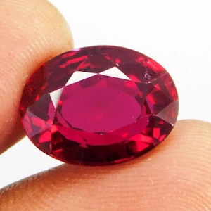 May include: A faceted, oval-shaped ruby gemstone. The gem is a deep, vibrant red color with multiple facets that reflect light. The ruby is held between two fingers, showcasing its size and brilliance.