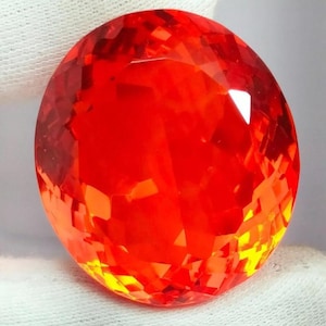 May include: A faceted, oval-shaped gemstone with a vibrant red-orange hue. The gem's surface reflects light, showcasing numerous facets that create a sparkling effect. The gemstone is likely a decorative item, suitable for jewelry or display.