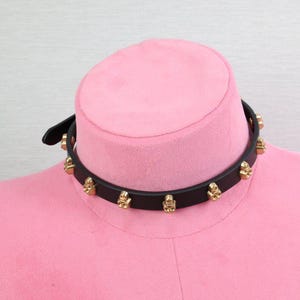 Puede incluir: Collar gargantilla negro con tachuelas de calavera doradas. El collar está hecho de una banda negra con adornos en forma de calavera espaciados uniformemente. La gargantilla se muestra en una cabeza de maniquí rosa.