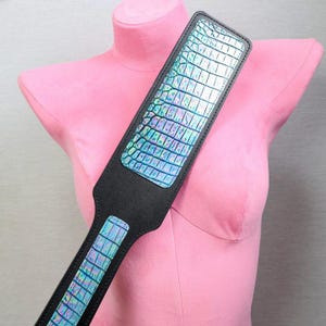 Puede incluir: Una paleta negra con un panel rectangular iridiscente con un patrón de piel de cocodrilo. La paleta se muestra contra un maniquí rosa, mostrando su diseño y textura. El mango también es negro.