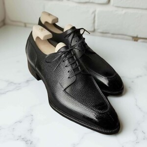 Puede incluir: Un par de zapatos de vestir de cuero negro con una parte superior texturizada y una puntera brillante. Los zapatos tienen cierre de cordones y se exhiben con hormas de madera en el interior. Sobre una superficie de m&aacute;rmol.