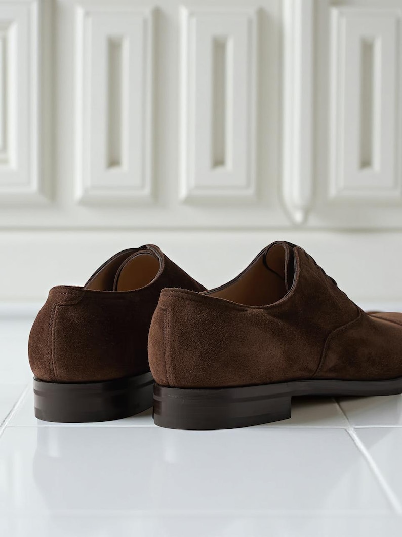 May include: Close-up of two brown suede dress shoes. The shoes have a classic Oxford design with a low heel and a dark brown sole. The suede material has a soft, textured appearance. The shoes are set against a white background.
