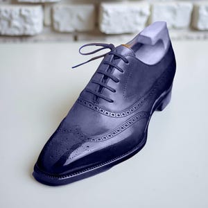 Pode incluir: Um sapato social de couro azul marinho com um design cl&aacute;ssico de biqueira. O sapato apresenta um acabamento brilhante, perfura&ccedil;&otilde;es intrincadas e fecho de atacadores. O sapato tem aproximadamente 30 cm de comprimento.