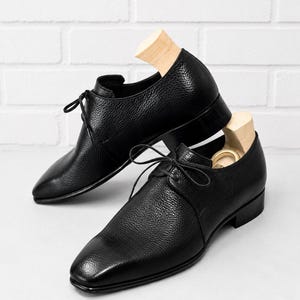 May include: A pair of black leather dress shoes with a textured finish, featuring a classic lace-up design. The shoes are displayed with wooden shoe trees inside, set against a white brick wall background.