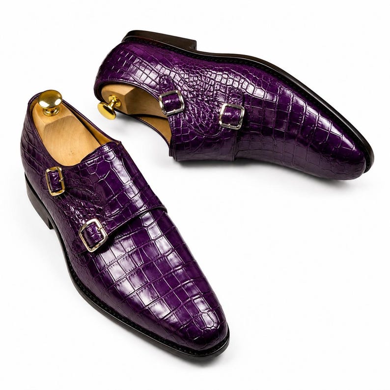 May include: A pair of purple leather double monk strap shoes with a crocodile texture. The shoes have gold-colored buckles and are set against a white background. The shoes are displayed with wooden shoe trees.