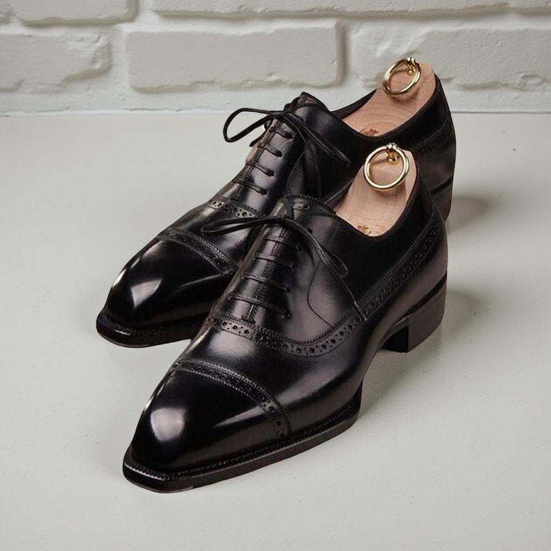May include: A pair of black leather dress shoes with a glossy finish. The shoes feature a classic Oxford design with decorative perforations and a lace-up closure. Shoe trees with gold-colored rings are inserted in the shoes.