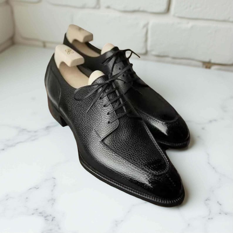 Puede incluir: Un par de zapatos de vestir de cuero negro con una parte superior texturizada y una puntera brillante. Los zapatos tienen cierre de cordones y se exhiben con hormas de madera en el interior. Sobre una superficie de m&aacute;rmol.