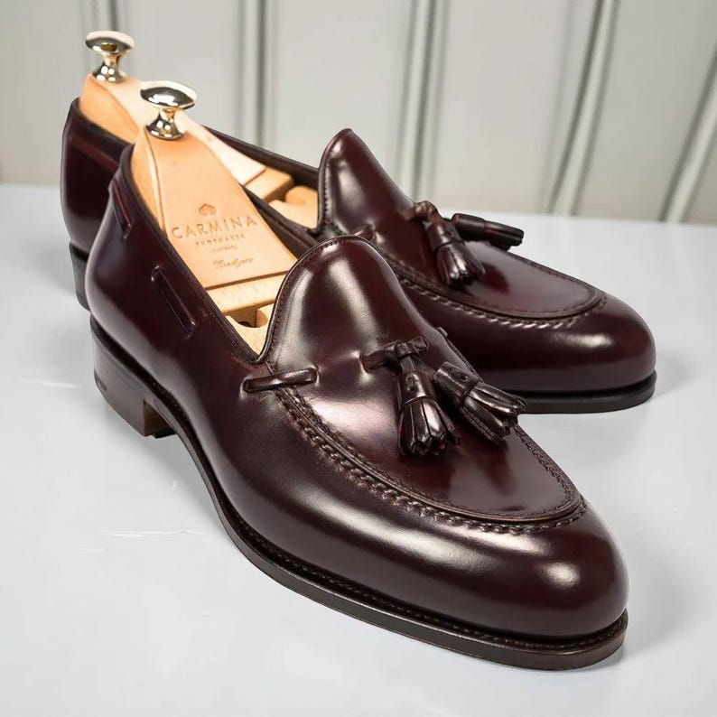May include: A pair of polished, burgundy leather loafers with decorative tassels. The shoes have a classic design with a rounded toe and a low heel. Shoe trees are inserted in the shoes. The brand name "CARMINA" is visible on the shoe tree.