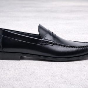 Op de afbeelding: Een enkele zwarte leren loafer schoen. De schoen heeft een klassiek penny loafer ontwerp met een ronde neus, een lage hak en zichtbare stikdetails. De schoen wordt gepresenteerd op een lichtgrijze ondergrond.