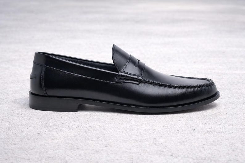 Op de afbeelding: Een enkele zwarte leren loafer schoen. De schoen heeft een klassiek penny loafer ontwerp met een ronde neus, een lage hak en zichtbare stikdetails. De schoen wordt gepresenteerd op een lichtgrijze ondergrond.