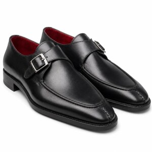 May include: A pair of black leather single monk strap shoes featuring a silver-toned metal buckle, a stitched apron toe design, and a vibrant red interior lining. The shoes have a low heel and a smooth, polished finish on the exterior surface.