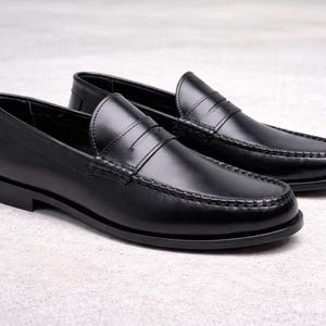Op de afbeelding: Een paar zwarte leren penny loafers. Deze klassieke instappers hebben een ronde neus, een band over de wreef en een lage hak. De schoenen worden gepresenteerd op een lichtgrijze ondergrond.