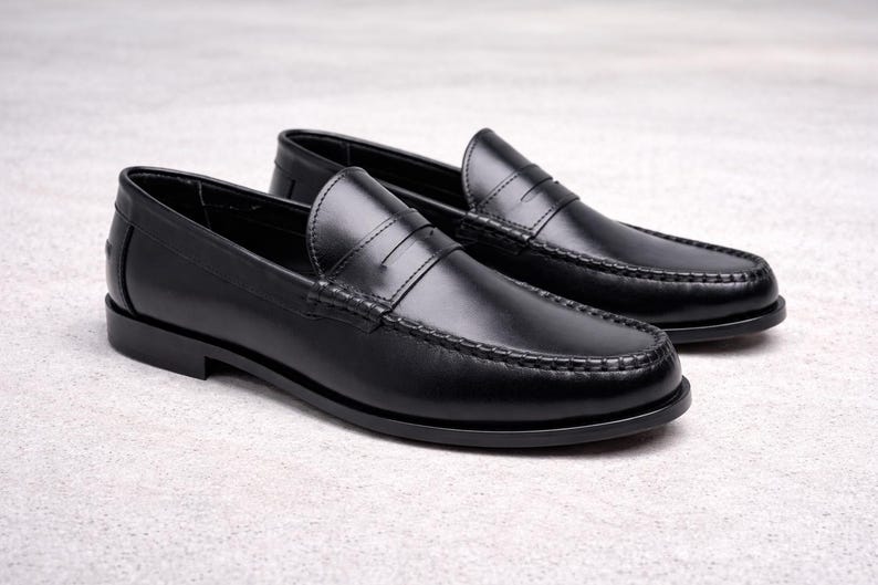 Op de afbeelding: Een paar zwarte leren penny loafers. Deze klassieke instappers hebben een ronde neus, een band over de wreef en een lage hak. De schoenen worden gepresenteerd op een lichtgrijze ondergrond.