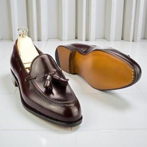 May include: A pair of polished, burgundy leather loafers with tassel details. One shoe is upright with a wooden shoe tree inside, the other is upside down, revealing a light brown sole. The shoes are on a white surface.