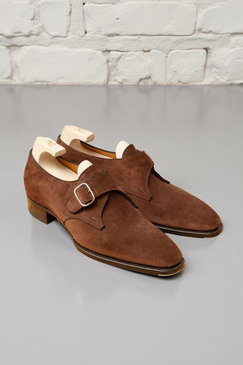 May include: A pair of brown suede dress shoes with a buckle closure. The shoes have a classic design with a rounded toe and a low heel. The shoes are displayed with wooden shoe trees inside.