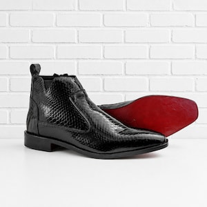 May include: A black ankle boot with a textured snakeskin pattern, a side zipper, and a pull tab at the heel. The boot features a low block heel and a smooth, vibrant red sole that is visible on the second boot positioned behind the first one.