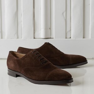 May include: A pair of brown suede Oxford shoes with a cap toe design. The shoes have a dark brown sole and laces. The background is a white wall with vertical paneling.