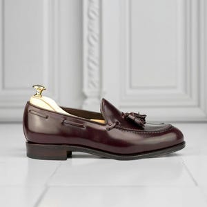 May include: A single, polished burgundy leather loafer with a tassel detail. The shoe has a rounded toe and a low heel. A wooden shoe tree is inside the shoe. The background is a white wall and floor.