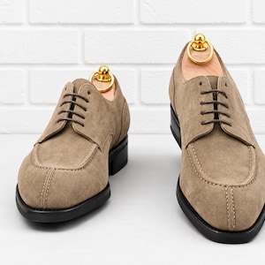 May include: Two suede dress shoes in a light brown color with black soles. The shoes have a classic design with a rounded toe and a lace-up closure. Shoe trees are inserted in the shoes to maintain their shape.