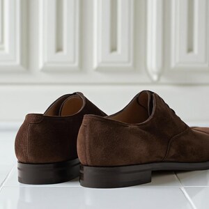 May include: Close-up of two brown suede dress shoes. The shoes have a classic Oxford design with a low heel and a dark brown sole. The suede material has a soft, textured appearance. The shoes are set against a white background.