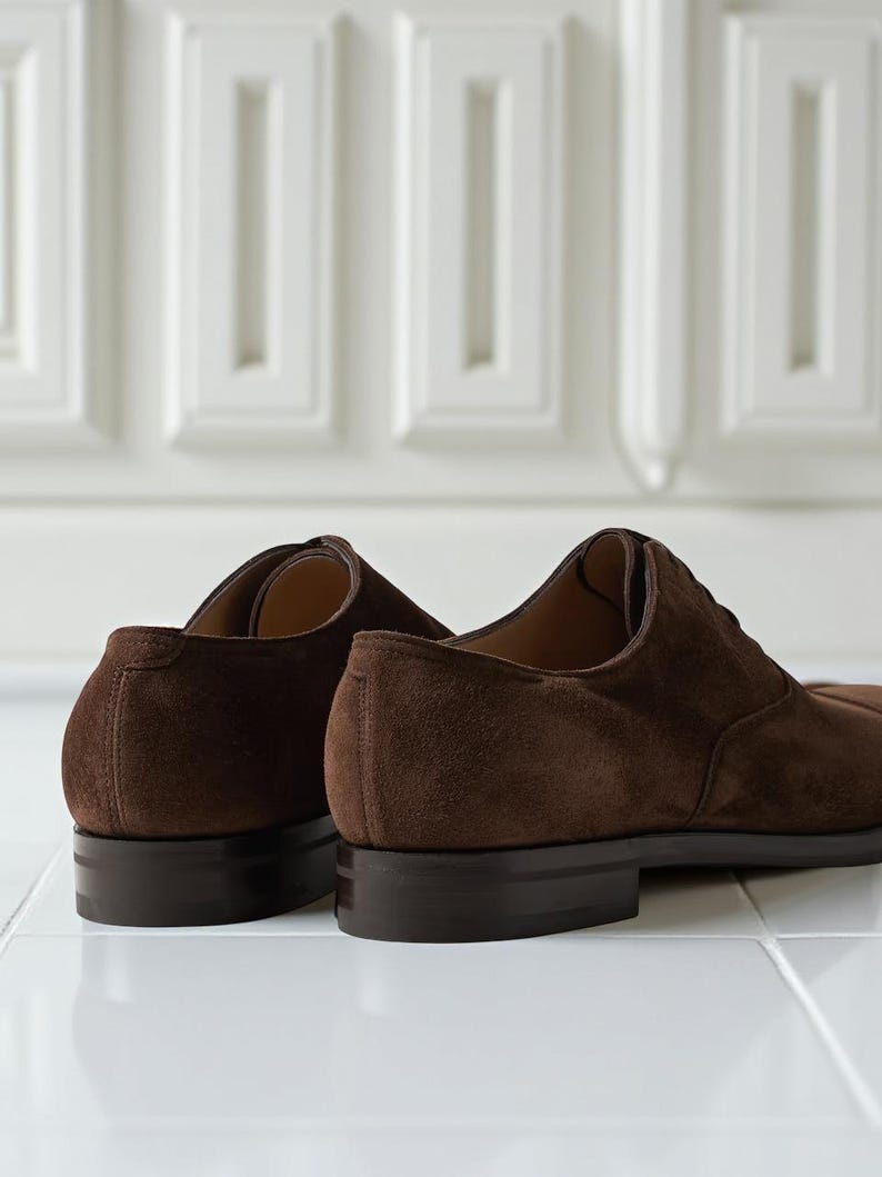 May include: Close-up of two brown suede dress shoes. The shoes have a classic Oxford design with a low heel and a dark brown sole. The suede material has a soft, textured appearance. The shoes are set against a white background.