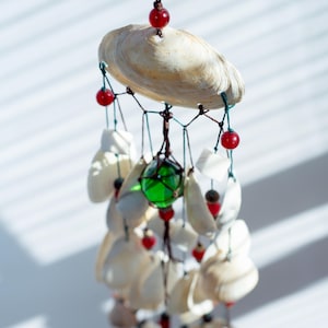 Può includere: Una carillon a vento di conchiglie con una grande conchiglia in cima, conchiglie più piccole appese sotto e perline rosse. Una palla di vetro verde è sospesa al centro. La carillon è sospesa da un cordoncino marrone.