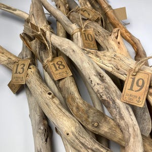 Peut inclure: Une collection de branches de bois flotté patinées dans des tons de beige, gris et marron. Chaque pièce est attachée par une ficelle à une étiquette en papier kraft portant le texte Sea Whisper Finds, une icône d'ancre et un numéro unique.