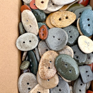 Può includere: Una collezione di bottoni in ceramica di forma ovale e irregolare in vari colori, tra cui beige, grigio, verde scuro e marrone rossastro. Ogni bottone presenta due fori per l'attacco e ha una finitura liscia, lucida o maculata.