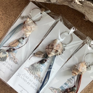 Puede incluir: Llaveros con conchas marinas, cuentas y detalles de cuero o tela. Cada llavero tiene un anillo de metal y una etiqueta que dice "FRIEND". El embalaje incluye el texto "LOST AT SEA, FOUND BY SHORE".