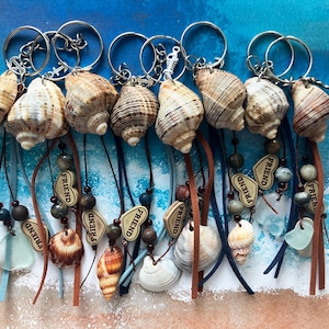 Op de afbeelding: Een verzameling sleutelhangers met zeeschelpen en zilverkleurige ringen. Elke sleutelhanger heeft een unieke zeeschelp, versierd met kralen, leren koorden en kleine houten bedels met de tekst "FRIEND". De achtergrond toont een strandscène.